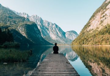 Man sitting on the end of a pier facing a beautiful lake surrounded by mountains